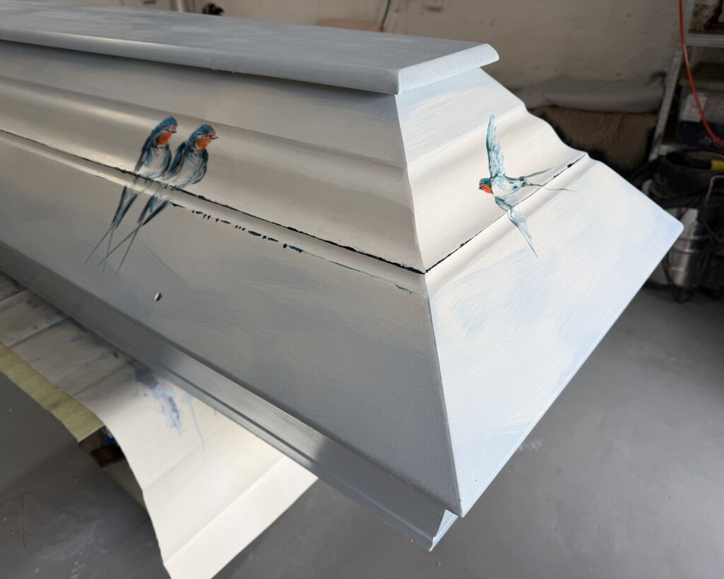 Ručně malovaná minimalistická rakev s motivem vlaštovek a jemnou morseovkou symbolizující naději, klid a návrat domů. Světle modro-bílá autorská malba Markéty Peškové pro důstojné poslední rozloučení.
